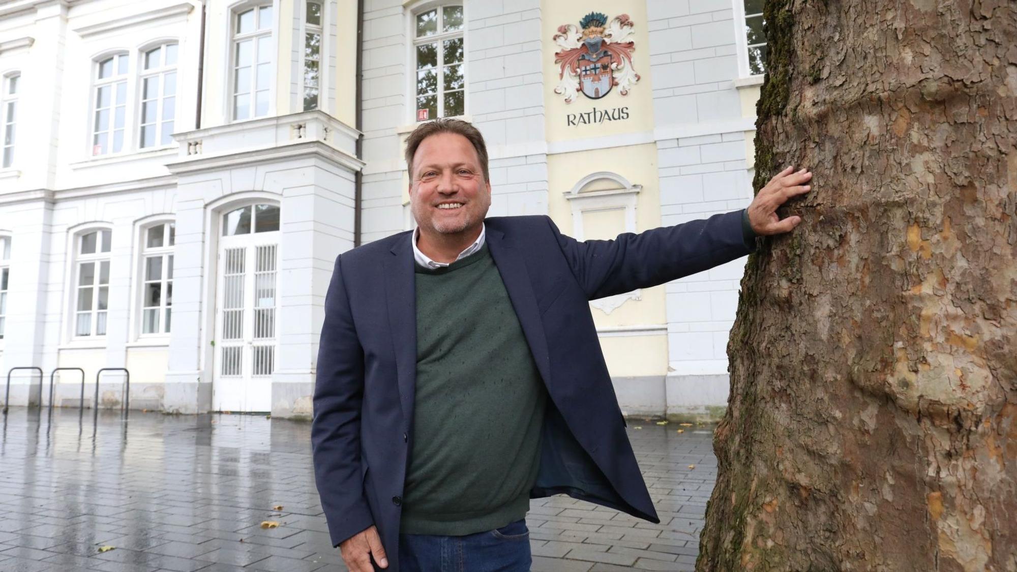 Ein Mann im blauen Sakko neben einem Baum, im Hintergrund an einer Fassade das Wappen der Stadt Königswinter und der Schriftzug Rathaus.