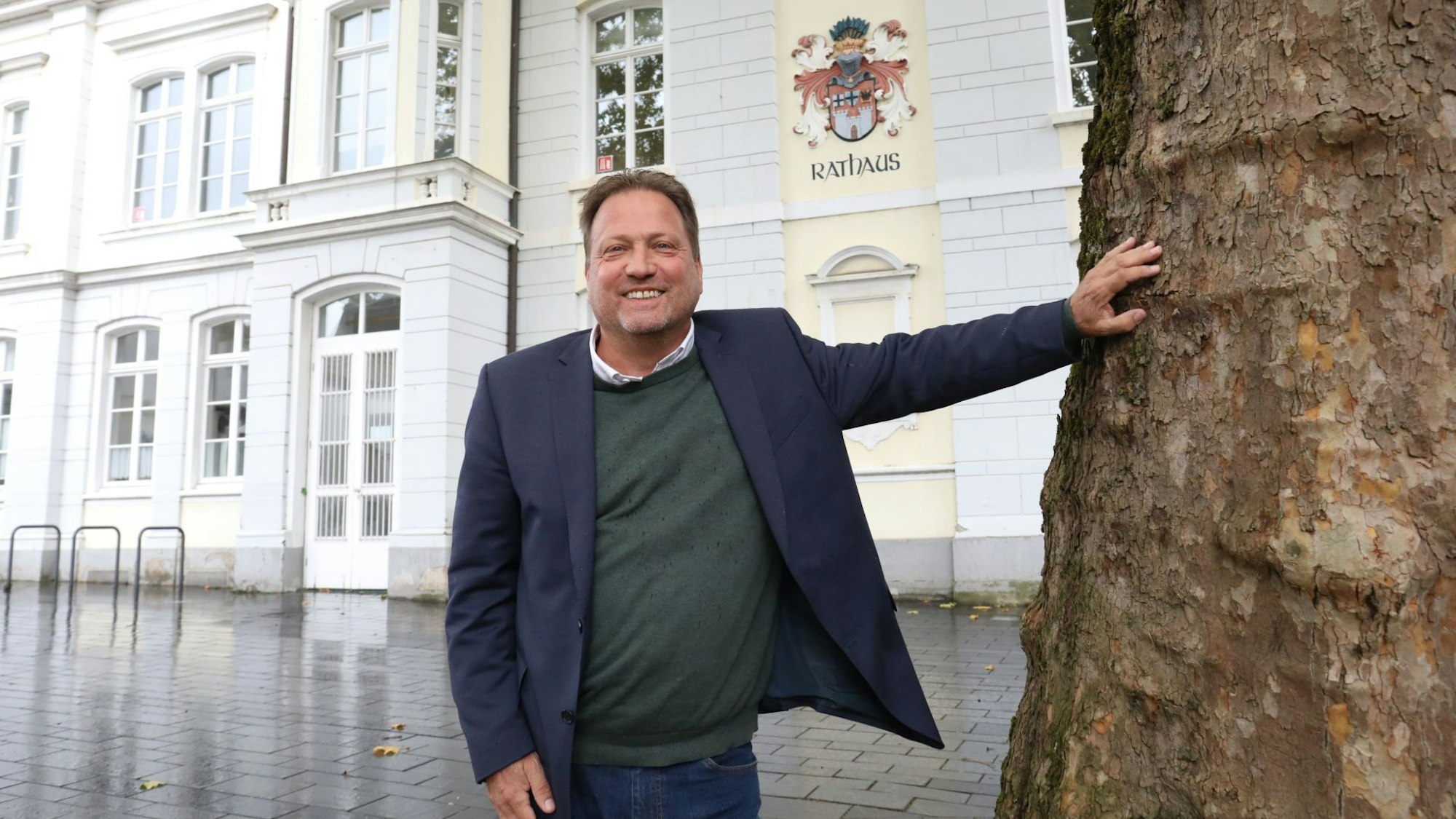 Ein Mann im blauen Sakko neben einem Baum, im Hintergrund an einer Fassade das Wappen der Stadt Königswinter und der Schriftzug Rathaus.