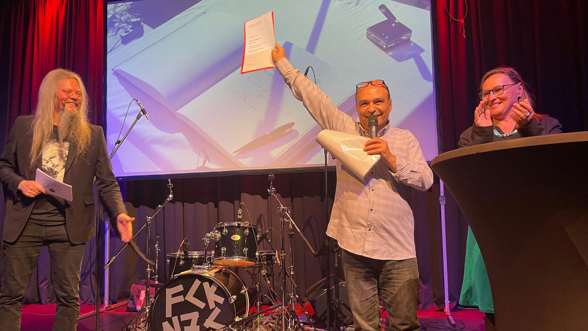 Ein Mann steht auf einer Bühne und hält lächelnd eine Urkunde hoch im Hintergrund steht Schlagzeug. neben ihm steht eine Frau und applaudiert.