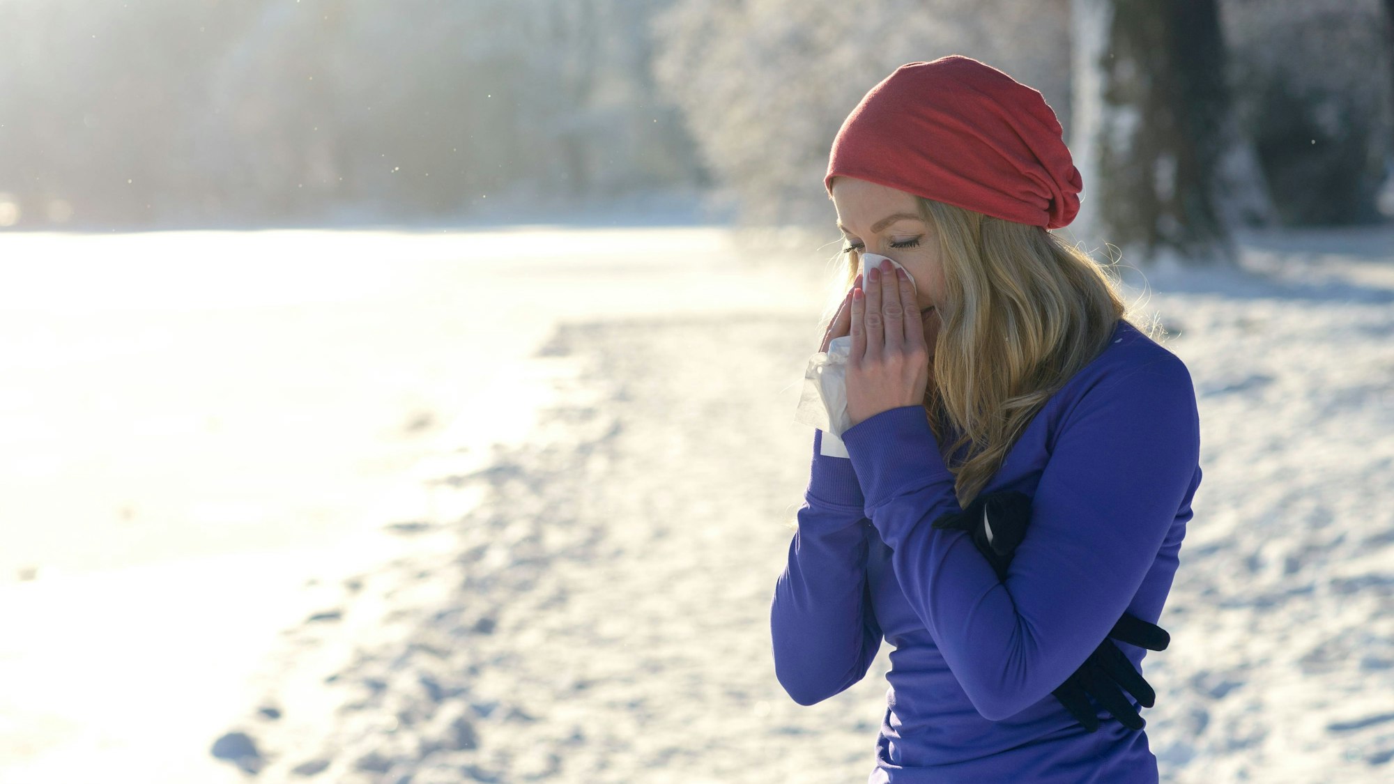 Eine erkältete Frau putzt sich auf einem Spaziergang durch den Schnee im Winter die Nase.