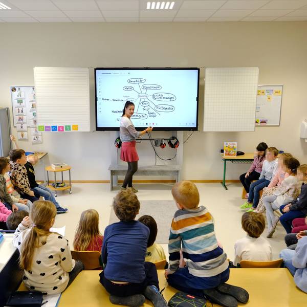 Blick in einen Klassenraum der Gemeinschaftsgrundschule Firmenich-Obergartzem. Die Lehrerin steht an der digitalen Tafel, die Schulkinder sitzen in einem Halbkreis davor.