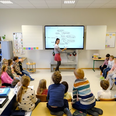Blick in einen Klassenraum der Gemeinschaftsgrundschule Firmenich-Obergartzem. Die Lehrerin steht an der digitalen Tafel, die Schulkinder sitzen in einem Halbkreis davor.