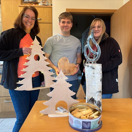 Michaela Lingscheid, Stefan Lingscheid und Ramona Bongartz stehen in einer Küche und halten hölzerne Dekorationsgegenstände in die Kamera.