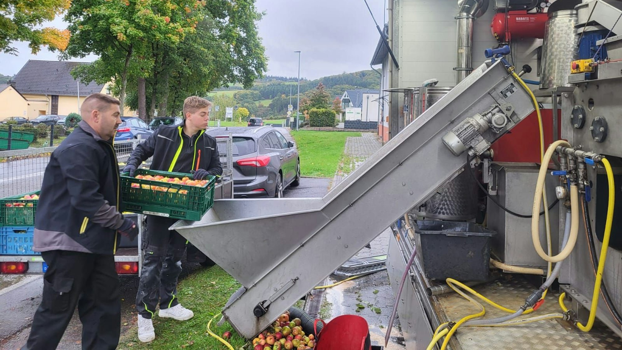 Peter und Stefan Lingscheid halten einen Korb voller Äpfel. Vater und Sohn setzen an, die Äpfel in eine Saftpresse zu kippen.