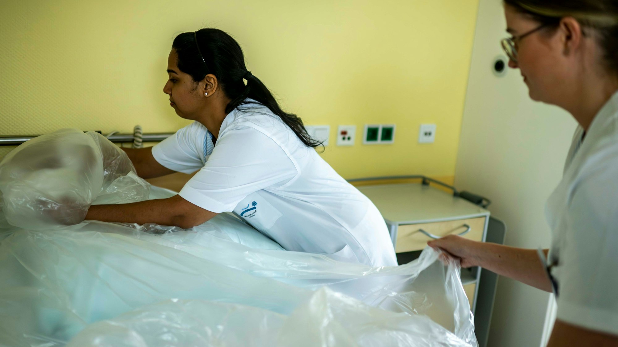Zwei Pflegefachkräfte des Euskirchener Marienhospitals bringen eine Plastikfolie an einem frisch bezogenen Bett an.