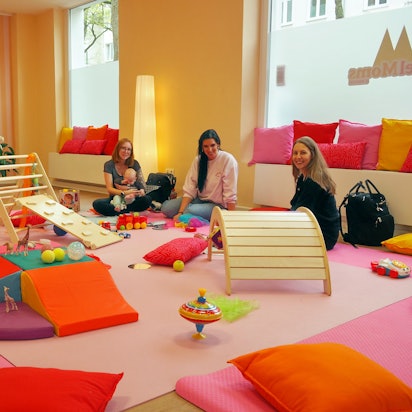 Drei Frauen sitzen in einem Raum voller bunter Matten und Spielgeräte für Kleinkinder.