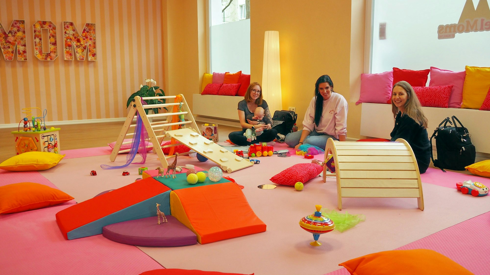 Drei Frauen sitzen in einem Raum voller bunter Matten und Spielgeräte für Kleinkinder.