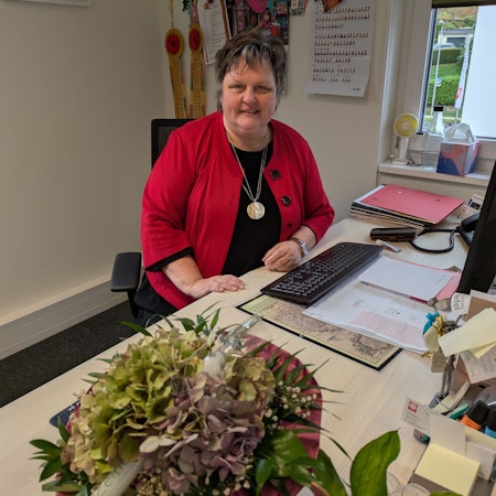 Ute Stolz sitzt am Schreibtisch, auf dem Unterlagen liegen und eine Vase mit Blumen steht.