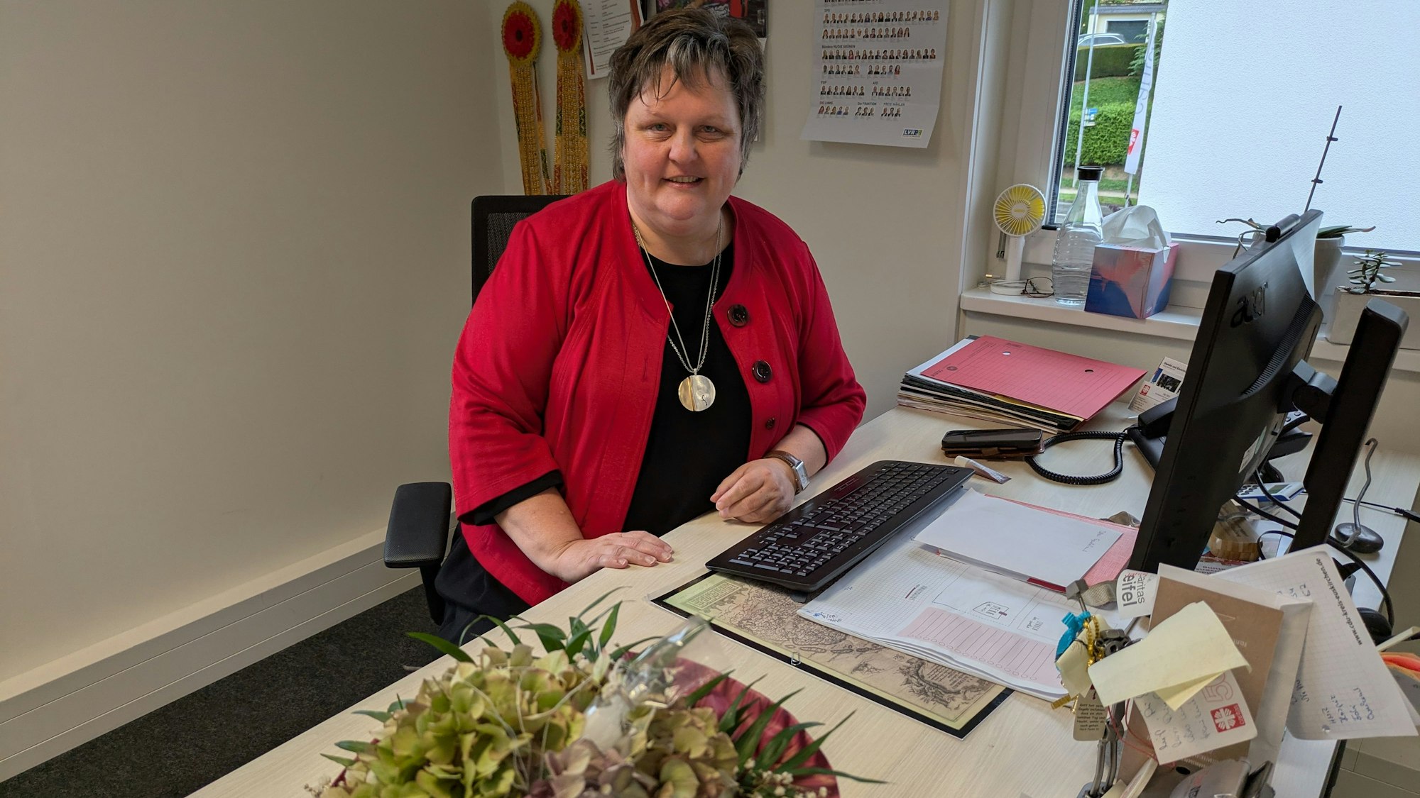 Ute Stolz sitzt am Schreibtisch, auf dem Unterlagen liegen und eine Vase mit Blumen steht.