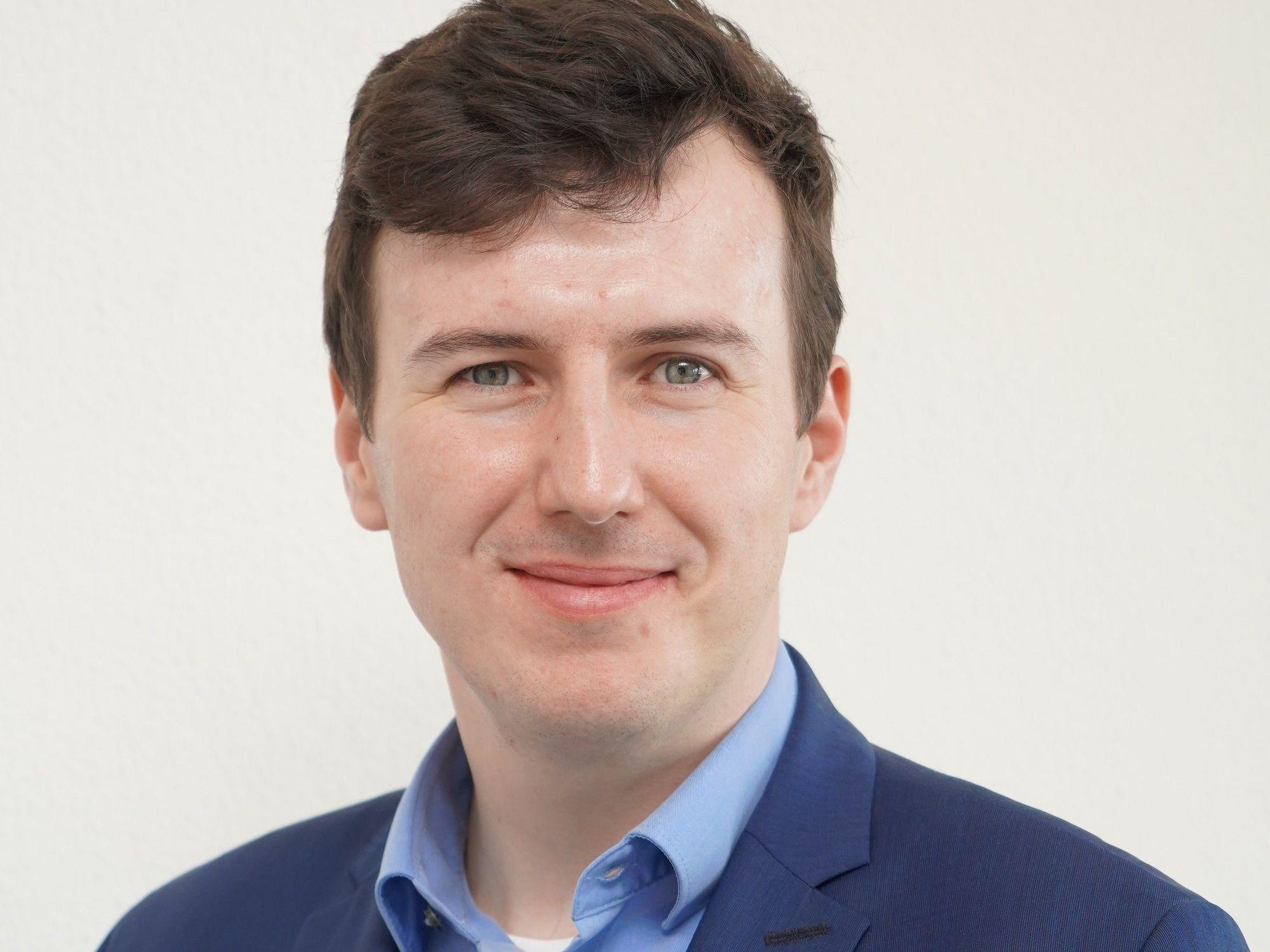 Auf dem Foto ist ein Portrait eines Mannes zu sehen, er trägt einen blauen Anzug und ein blaues Hemd.