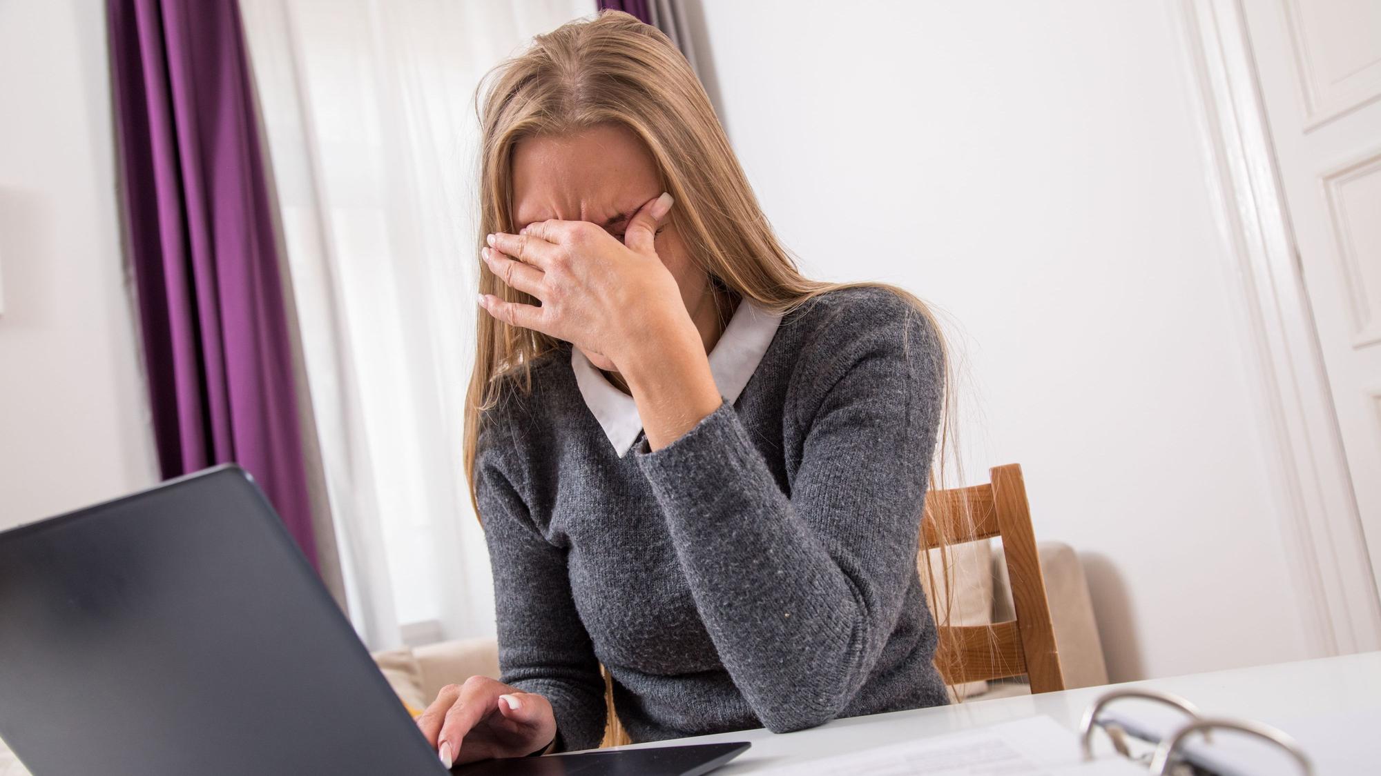 Eine Frau sitzt vor ihrem Laptop und hält die Hand vor Augen, weil sie Kopfschmerzen hat.