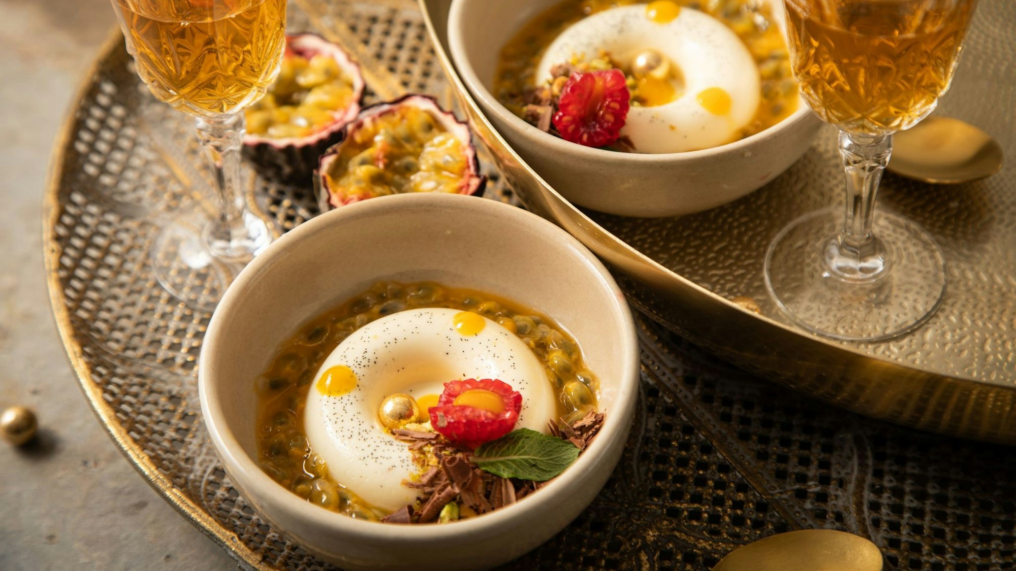 Panna Cotta mit Passionsfrucht, Himbeere, Mangotupfer, Minze, Pistazie, Schokolade und goldenen Schokokugeln in Savarin-Form zubereitet. Das ist eine Backform in flacher, ringförmiger Gestalt.