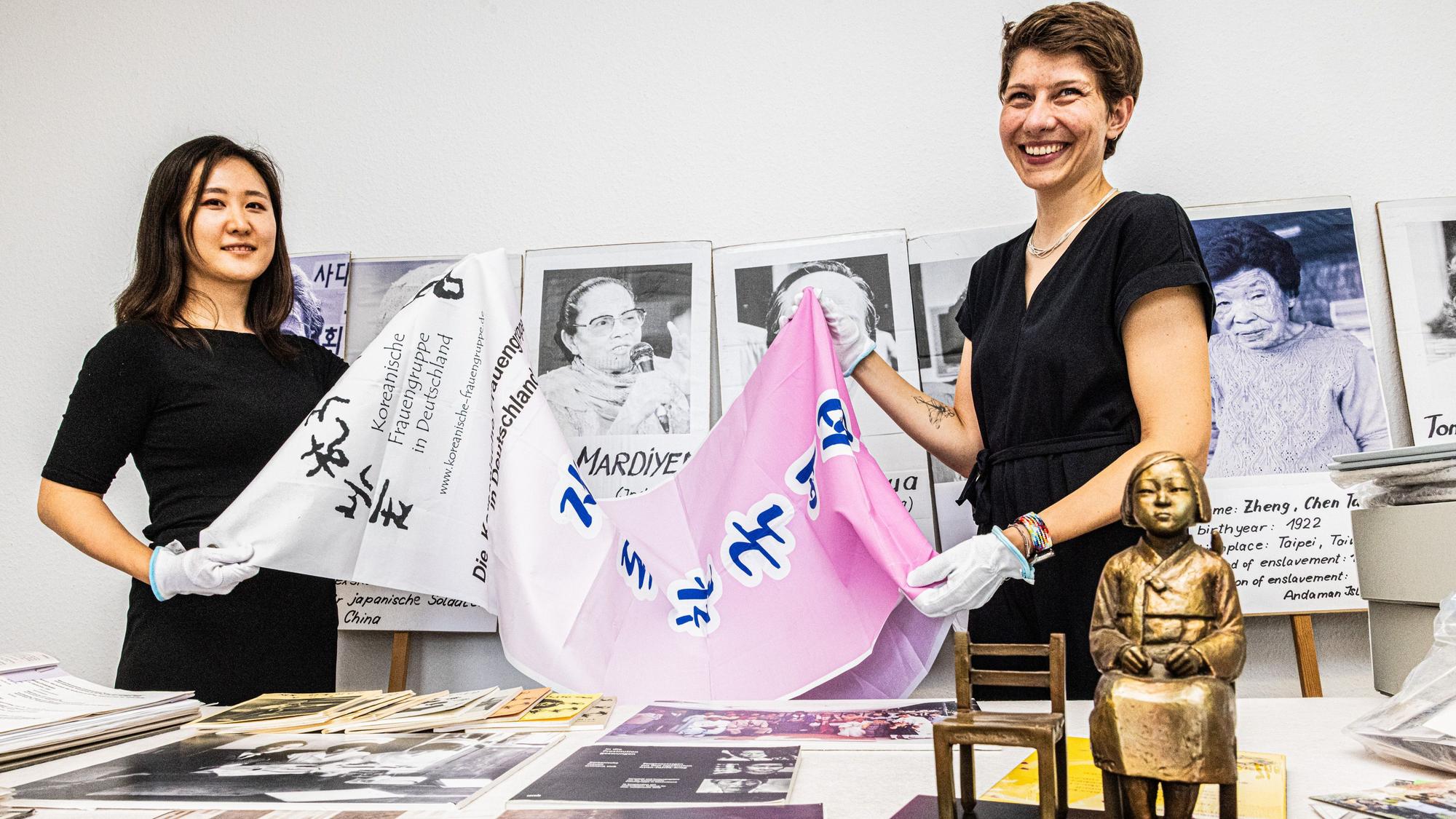 Ein Banner der koreanischen Frauengruppe zeigen Hojin Lee und Vera Tönsfeldt (v.l.). Dahinter sind Stabplakate mit Gesichtern von Frauen zu sehen, die von Militärs vergewaltigt wurden. Damit beteiligten sich die Vereinsfrauen an Protesten.