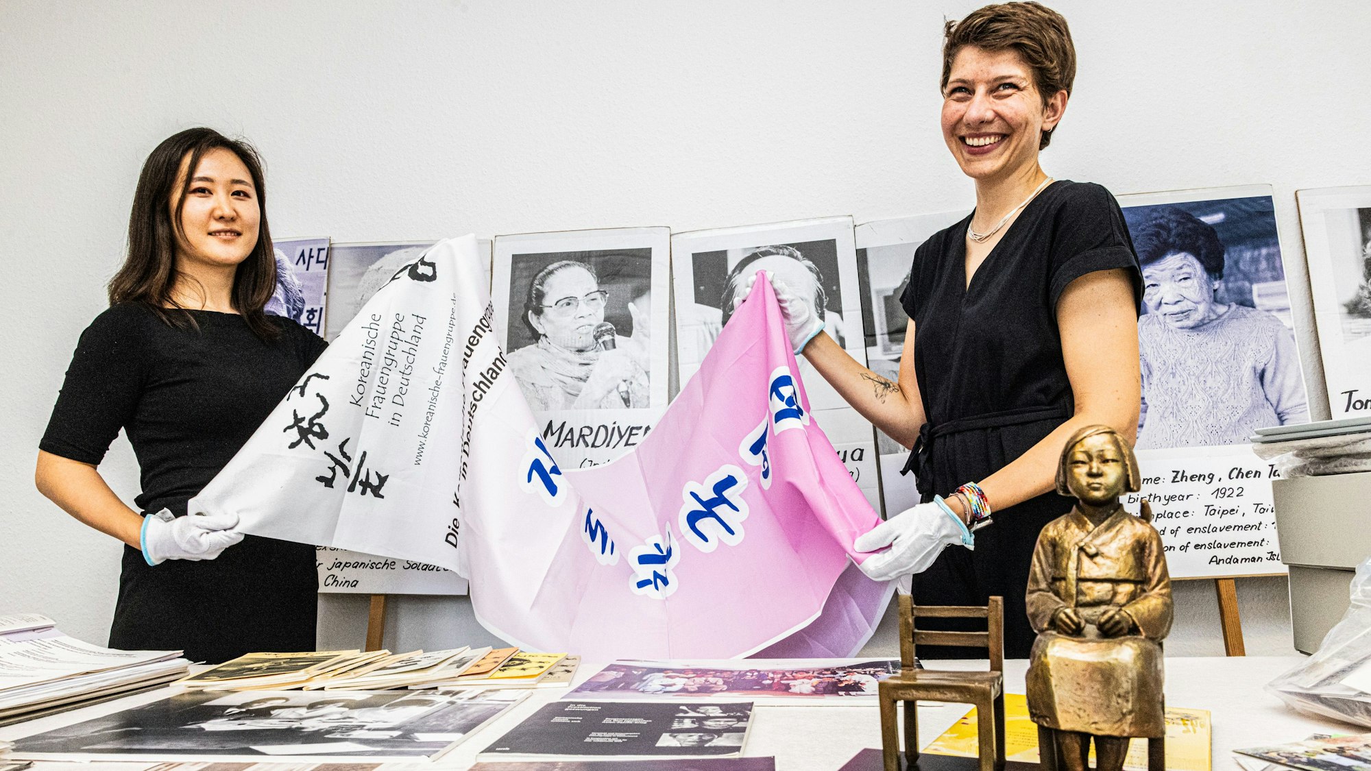 Ein Banner der koreanischen Frauengruppe zeigen Hojin Lee und Vera Tönsfeldt (v.l.). Dahinter sind Stabplakate mit Gesichtern von Frauen zu sehen, die von Militärs vergewaltigt wurden. Damit beteiligten sich die Vereinsfrauen an Protesten.