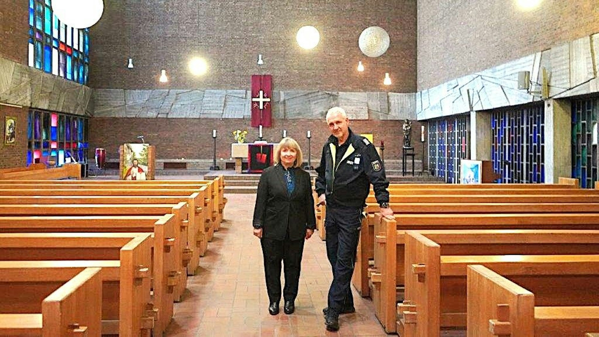 Eine blonde Frau steht neben einem großen Mann in Uniform in einem Kirchenraum zwischen zwei parallelen Bankreihen.