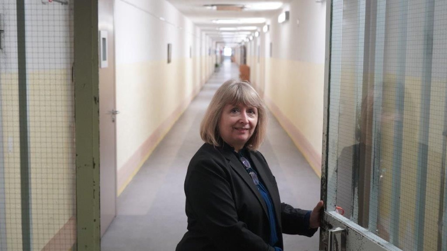 Eine Frau mit blondem schulterlangem Haar steht an einer Tür, hinter der ein langer Gang liegt.