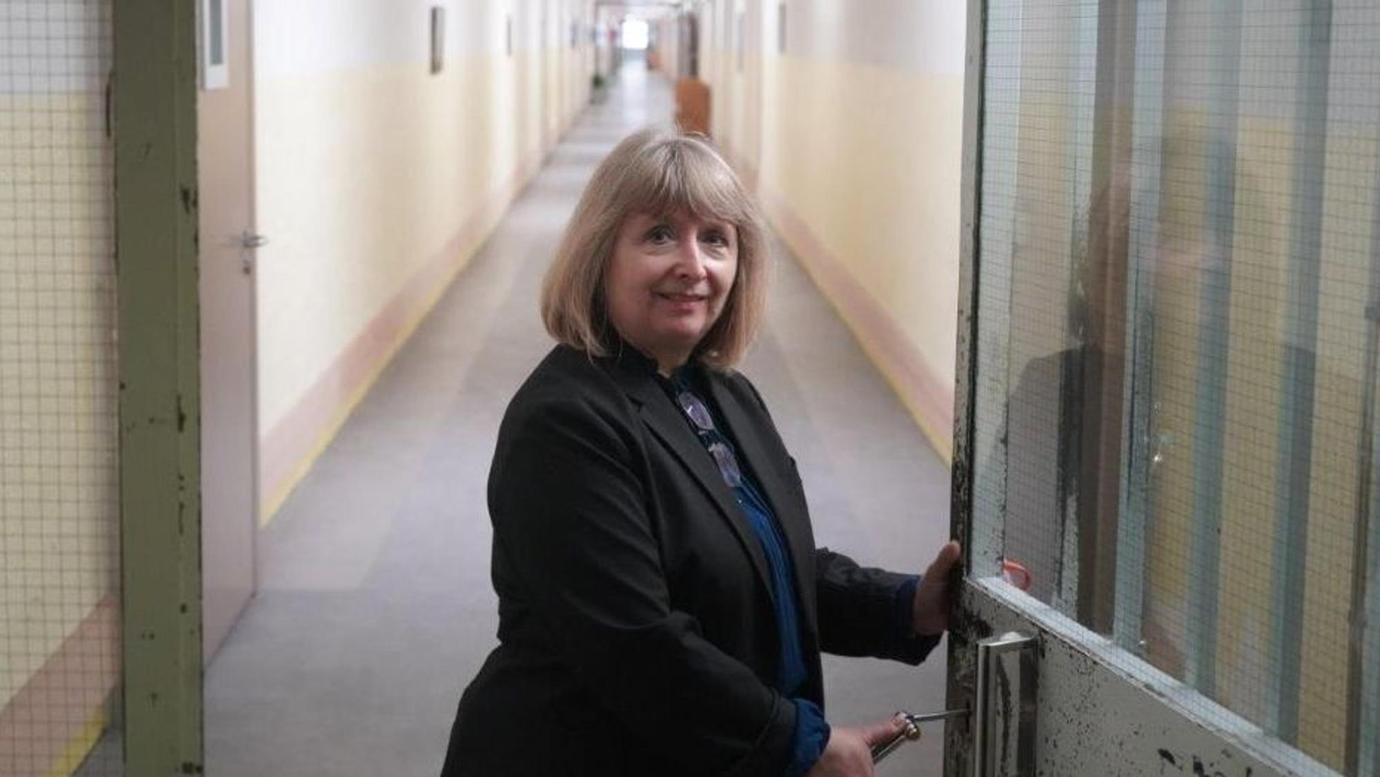 Eine Frau mit blondem schulterlangem Haar steht an einer Tür, hinter der ein langer Gang liegt.