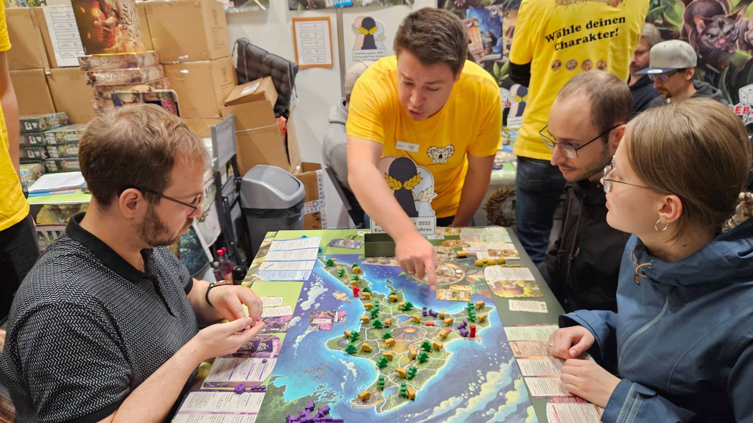 Drei junge Leute sitzen an einem Brettspiel, das eine Insel zeigt. Ein Mann im gelben T-Shirt erklärt die Regeln.