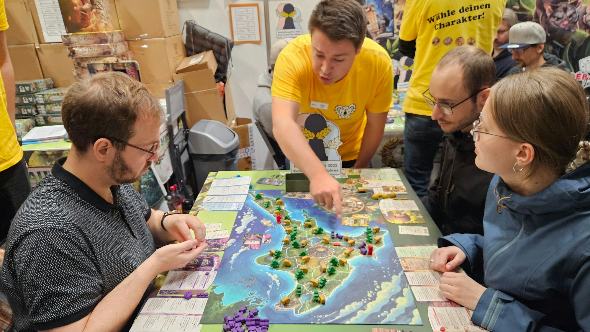 Drei junge Leute sitzen an einem Brettspiel, das eine Insel zeigt. Ein Mann im gelben T-Shirt erklärt die Regeln.
