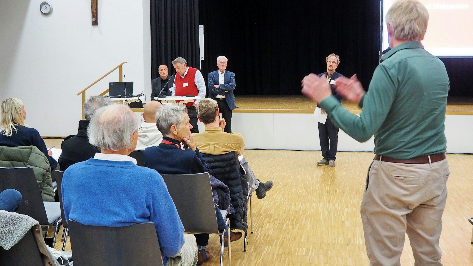 Menschen sitzen auf Stühlen in einer Schul-Aula.