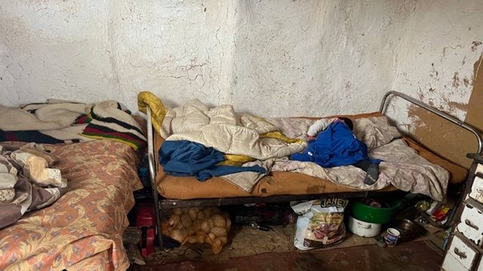 Zwei einfache Betten stehen in einer L-Form zueinander in einem Raum, an dem die Farbe von der Wand bröckelt. Die Matratzen und Bettwäsche sieht alt und schmutzig aus. Essensvorräte sind unter den Betten gelagert.
