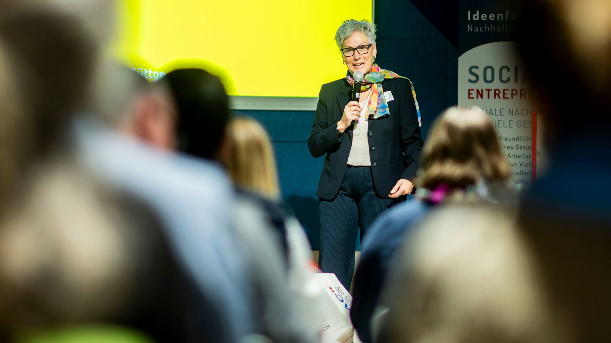 Iris Poth steht vor einer Gruppe Menschen, deren Hinterköpfe sich unscharf auf dem Foto befinden.