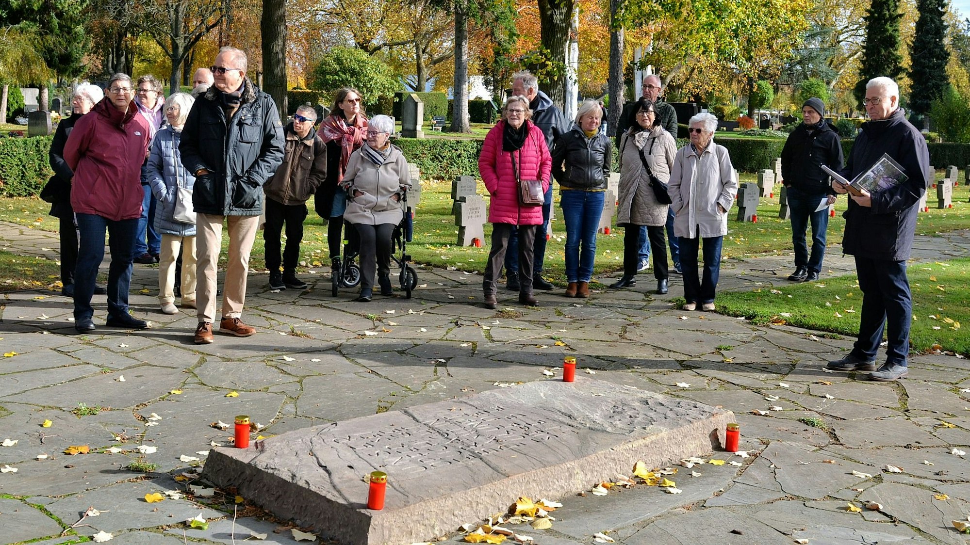 Eine Besuchergruppe steht vor einem Grab, dessen waagerechte Steintafel mit vier an den Ecken aufgestellten Kerzen geschmückt ist. Kurt Lingscheidt liest aus einem Buch vor.
