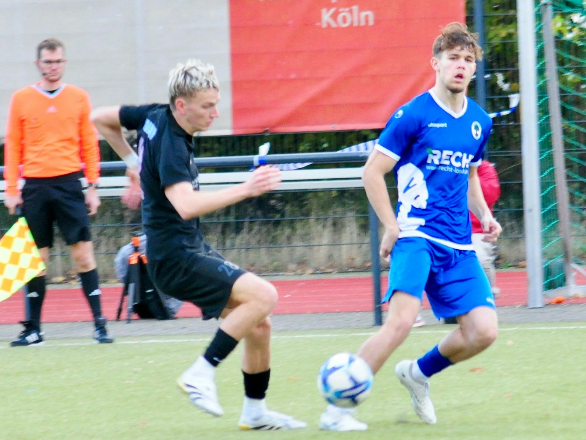 Das Foto zeigt einen Fußballer im blauen Trikot, der den Ball vor seinem heraneilenden Gegenspieler in die Mitte des Spielfelds passt.