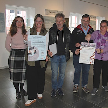 Sieben Personen stehen nebeneinander und lächeln in die Kamera. In den Händen halten sie Plakate, die für die Veranstaltungen werben.