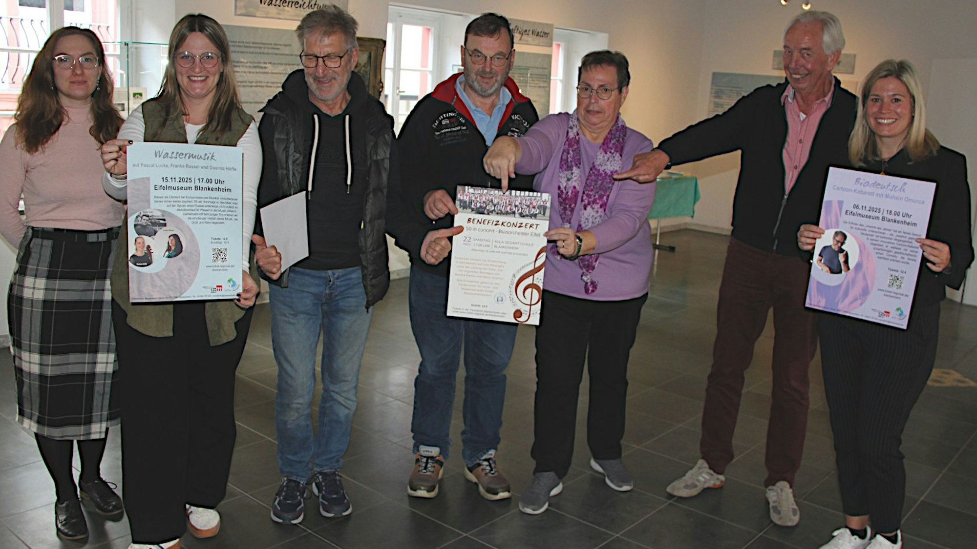 Sieben Personen stehen nebeneinander und lächeln in die Kamera. In den Händen halten sie Plakate, die für die Veranstaltungen werben.