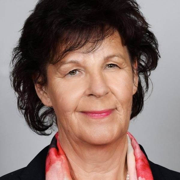Eine Frau mit dunklem Haar, dunklem Blazer und Perlenkette schaut lächelnd in die Kamera.