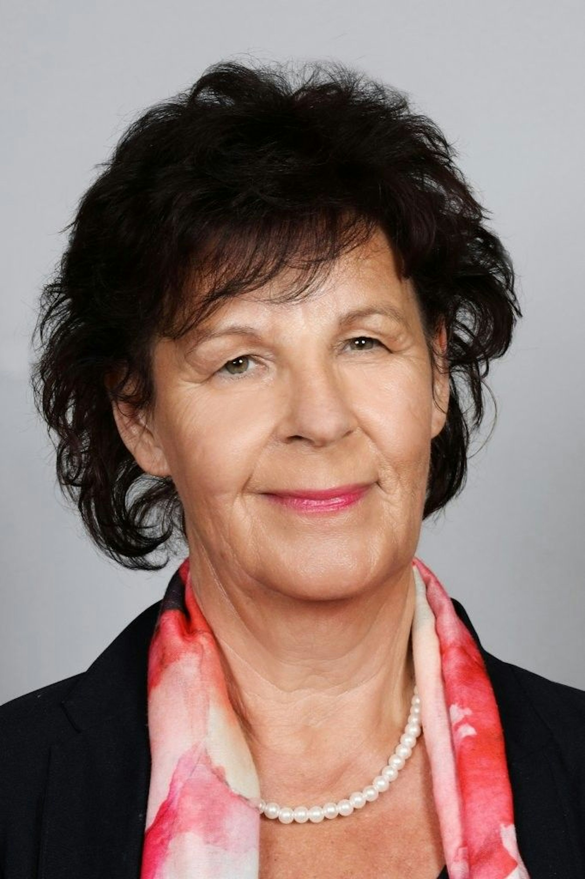 Eine Frau mit dunklem Haar, dunklem Blazer und Perlenkette schaut lächelnd in die Kamera.