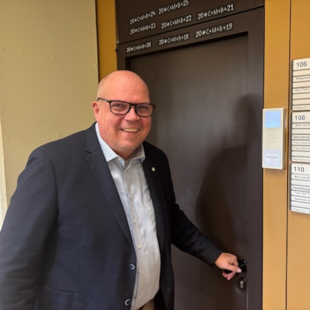 Auf dem Bild ist ein Mann im blauen Sakko zu sehen, der eine braune Bürotür öffnet.