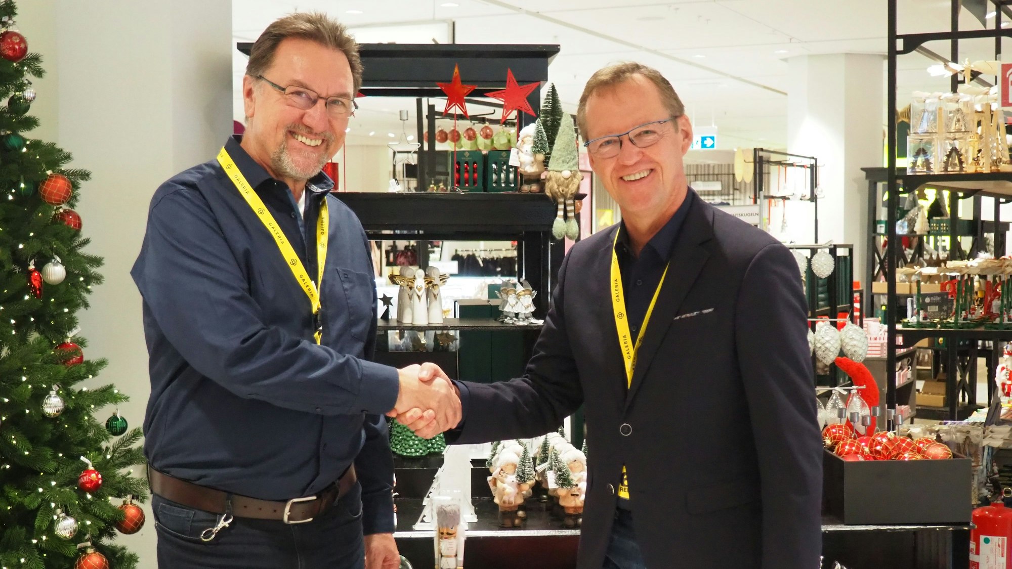 Harry Benzrath (l.) und Gerhard Ordczinsky schütteln sich in einem Galeria Geschäft, das weihnachtlich dekoriert ist, die Hände.