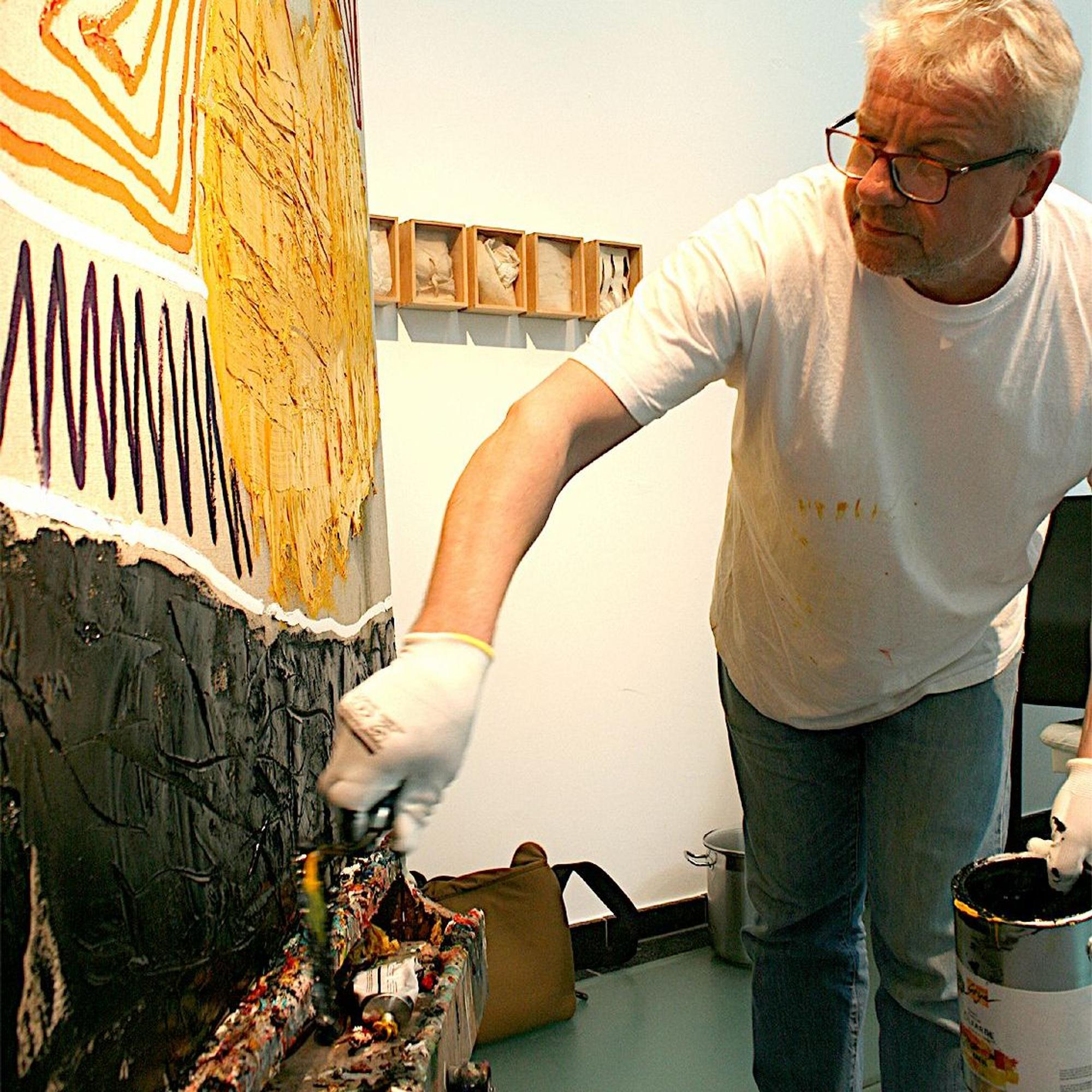Der Künstler steht an einer Leinwand, in der Hand hält er einen Eimer Farbe, mit der anderen spachtelt er eine Schicht schwarze Farbe auf.