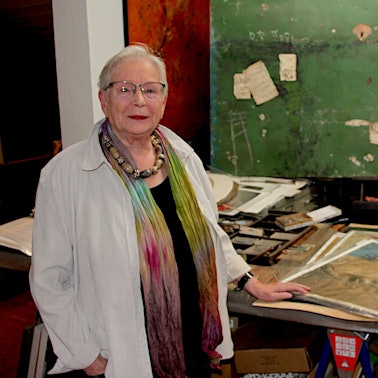 Eva-Maria Hermanns steht im Atelier, vor ihr liegen lose Blätter mit Malereien.