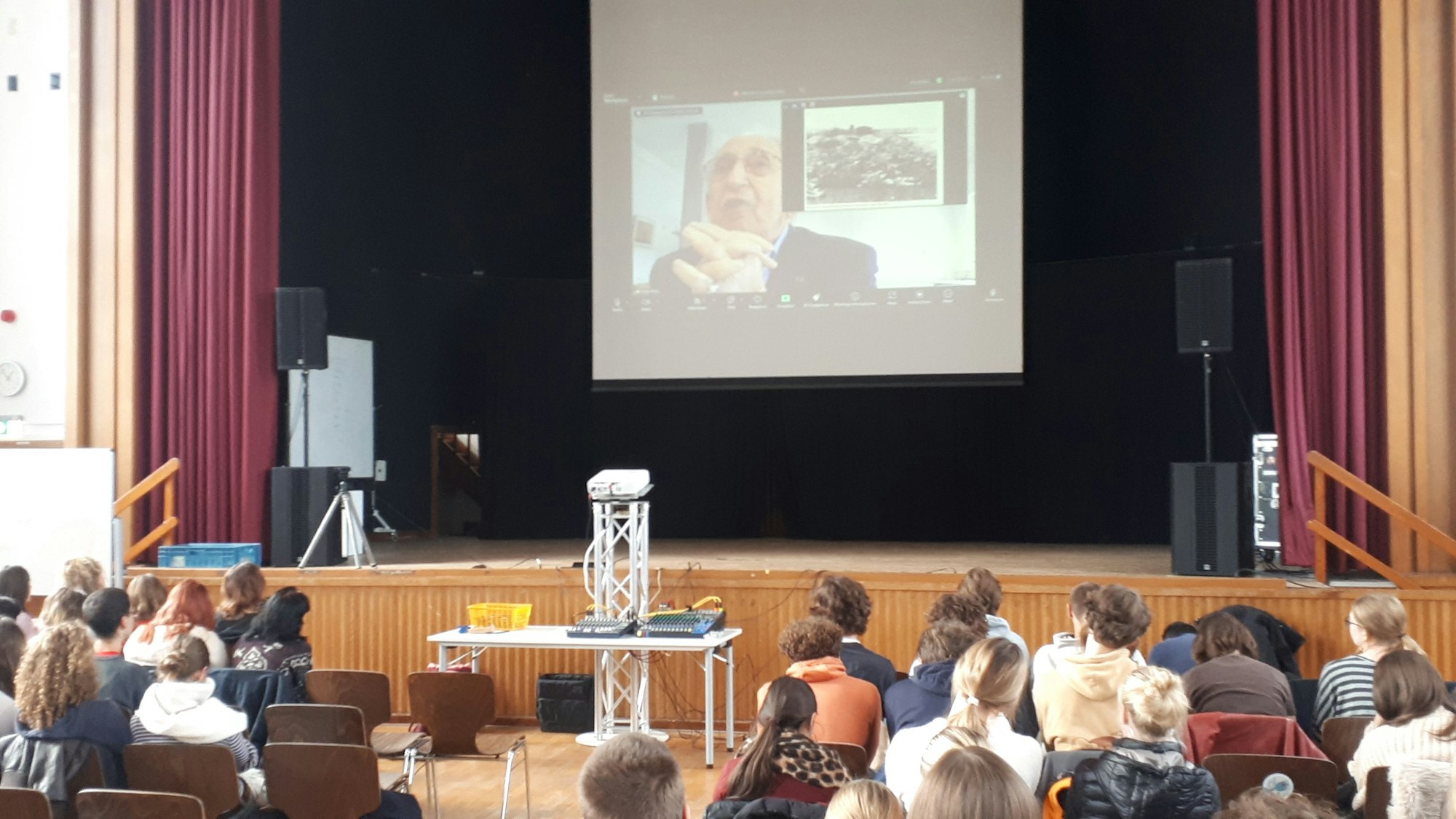 In einer Schulaula hängt über der Bühne eine Leinwand, auf der ein alter Mann zu sehen ist. Er beantwortet Fragen der Schüler, die in der Aula sitzen.