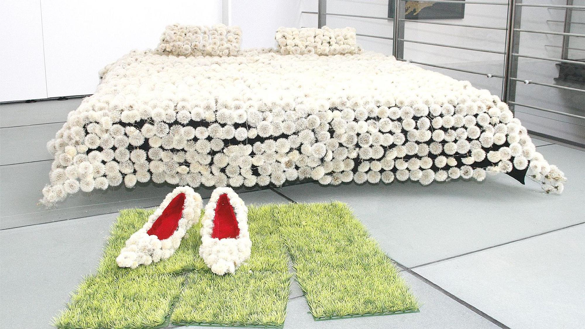 Die Installation zeigt ein Bett mit einer Pusteblumen-Decke, davor liegen Rasenstücke, auf denen überdimensionierte Pumps stehen, ebenfalls aus Pusteblumen.