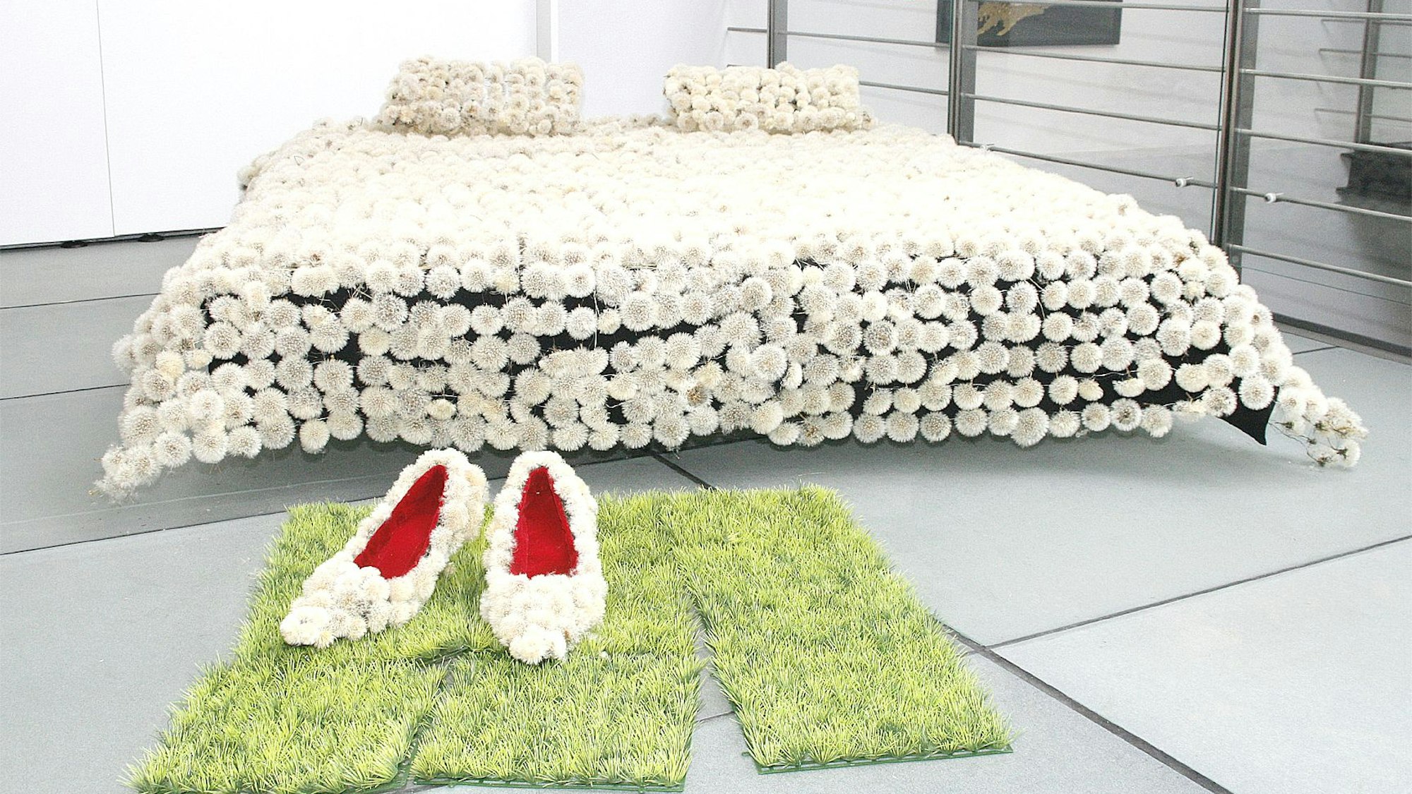 Die Installation zeigt ein Bett mit einer Pusteblumen-Decke, davor liegen Rasenstücke, auf denen überdimensionierte Pumps stehen, ebenfalls aus Pusteblumen.