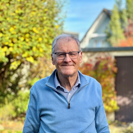 Ein Mann mit Brille, blauem Pullover und grauen Haaren steht in einem Garten.