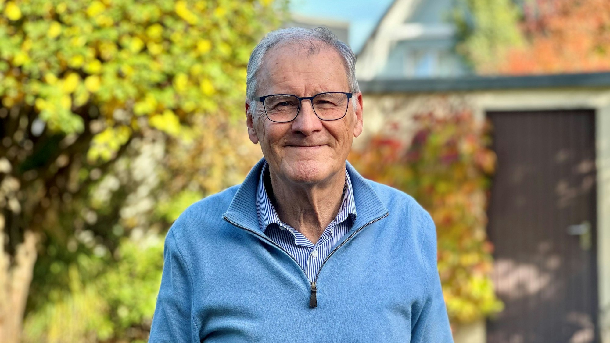Ein Mann mit Brille, blauem Pullover und grauen Haaren steht in einem Garten.