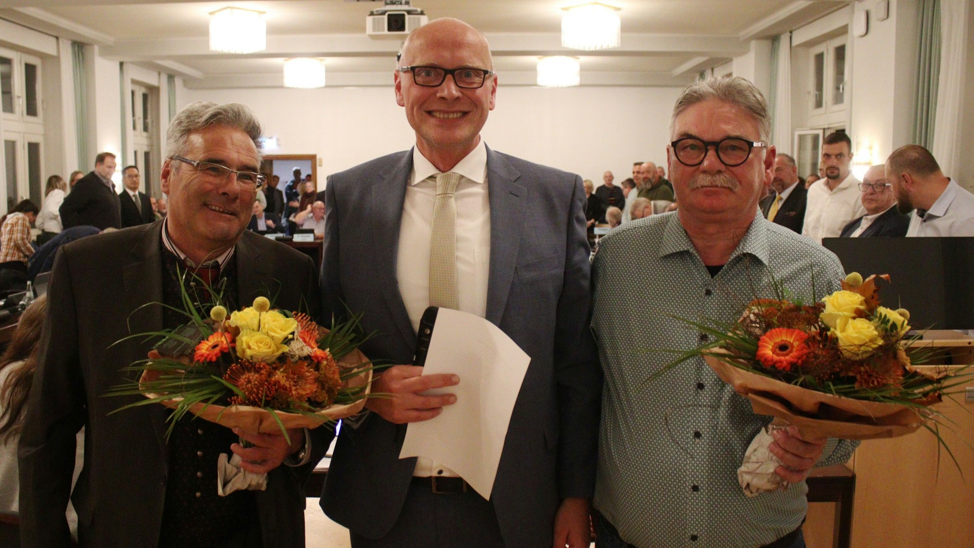 Rechts und links vom Bürgermeister stehen dessen Stellvertreter, die beide Blumen in den Händen halten.