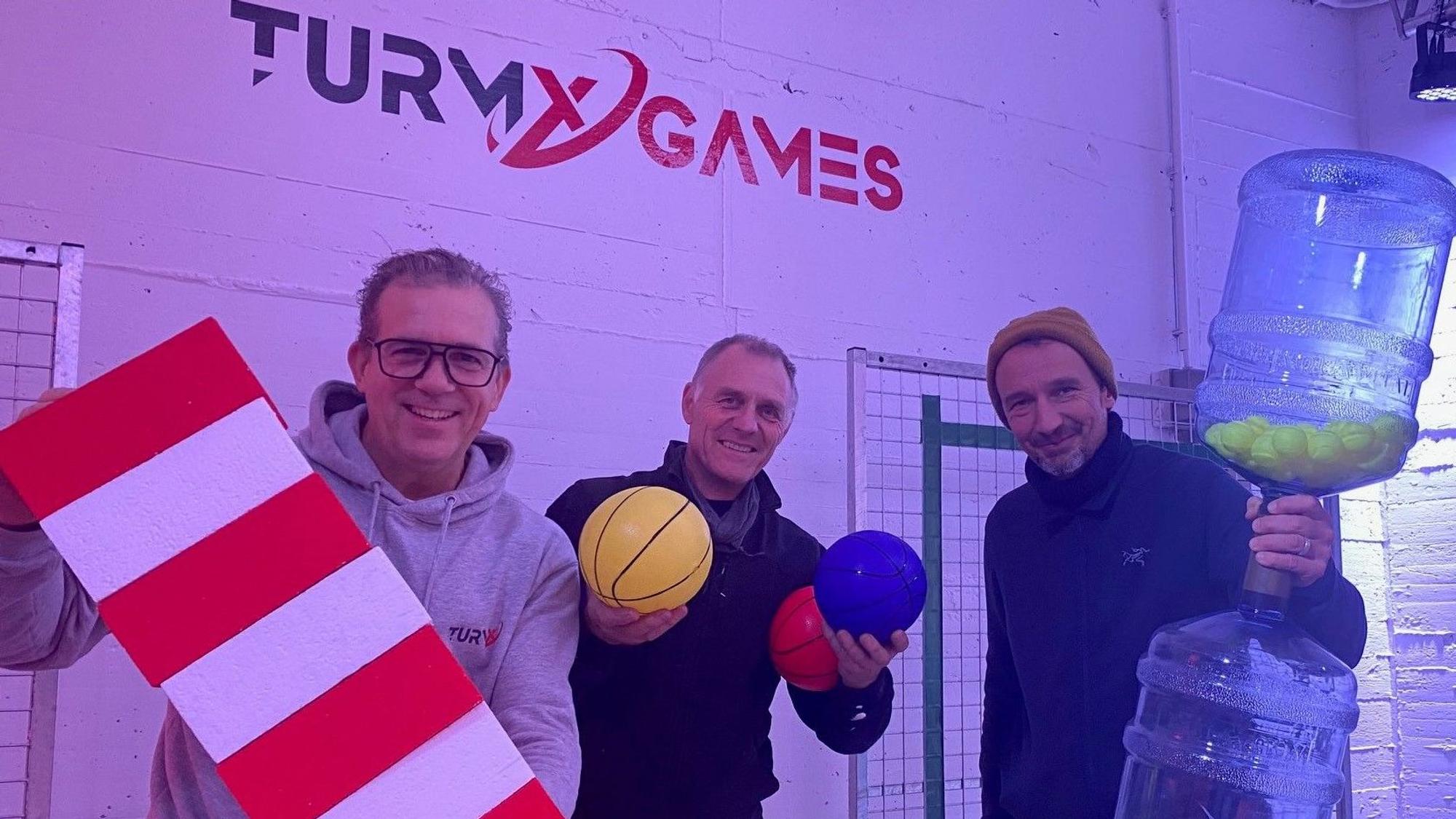 Zu sehen sind Achim Heymann, Marco Gleißner und Raimund Bechtloff, sie halten Gameshow-Equipment in den Händen.