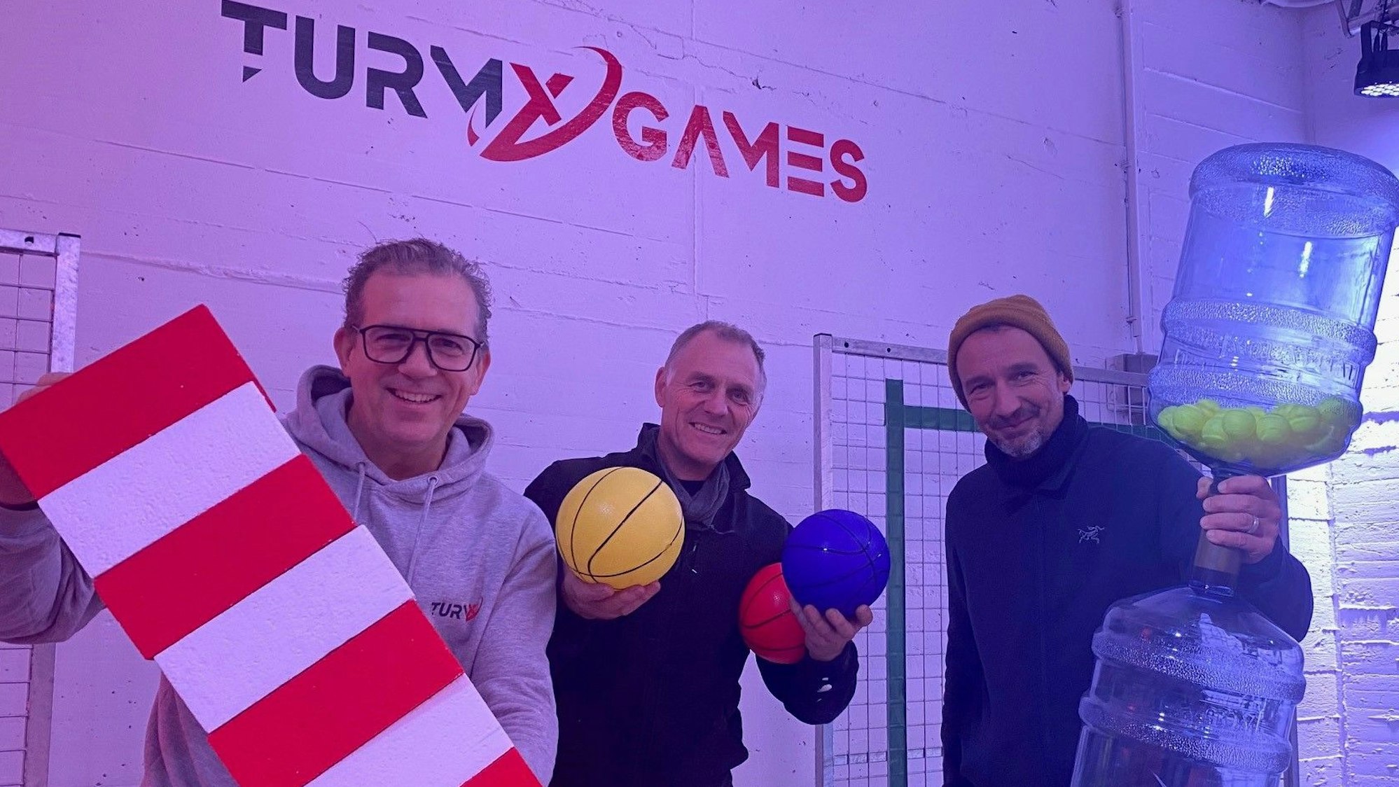 Zu sehen sind Achim Heymann, Marco Gleißner und Raimund Bechtloff, sie halten Gameshow-Equipment in den Händen.