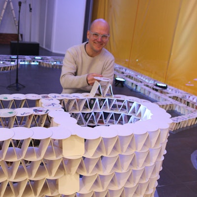 Ein Mann kniet hinter einem Bauwerk, das schon jetzt aus Tausenden Bierdeckeln.