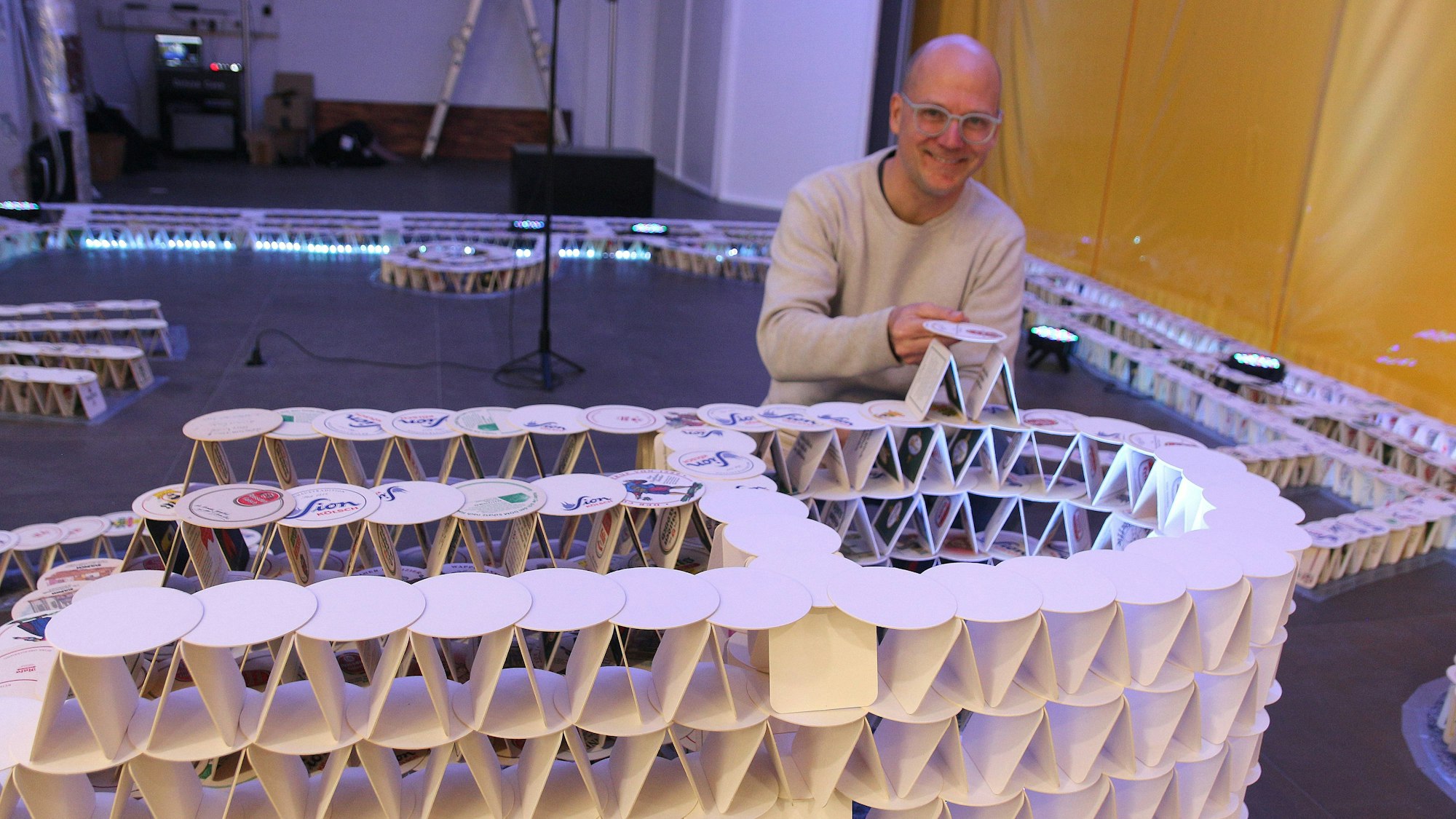 Ein Mann kniet hinter einem Bauwerk, das schon jetzt aus Tausenden Bierdeckeln.