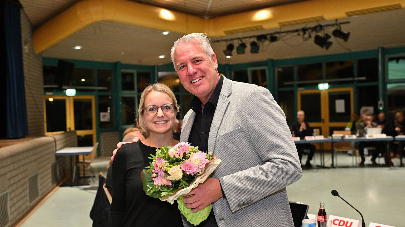 Eine Frau und in Mann stehen in einem Ratssaal und umarmen sich. Er hält einen Blumenstrauß in der Hand.