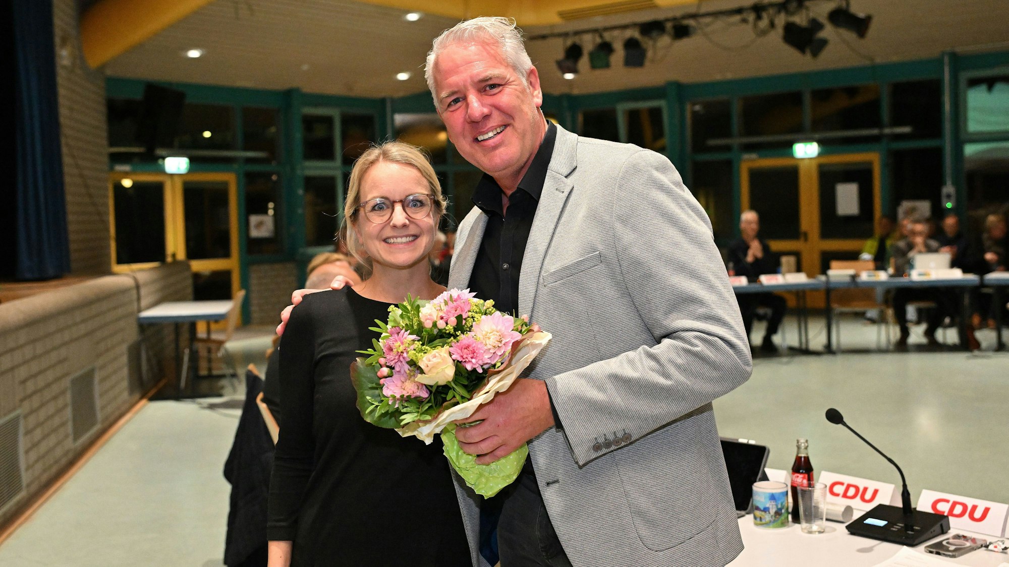 Eine Frau und in Mann stehen in einem Ratssaal und umarmen sich. Er hält einen Blumenstrauß in der Hand.