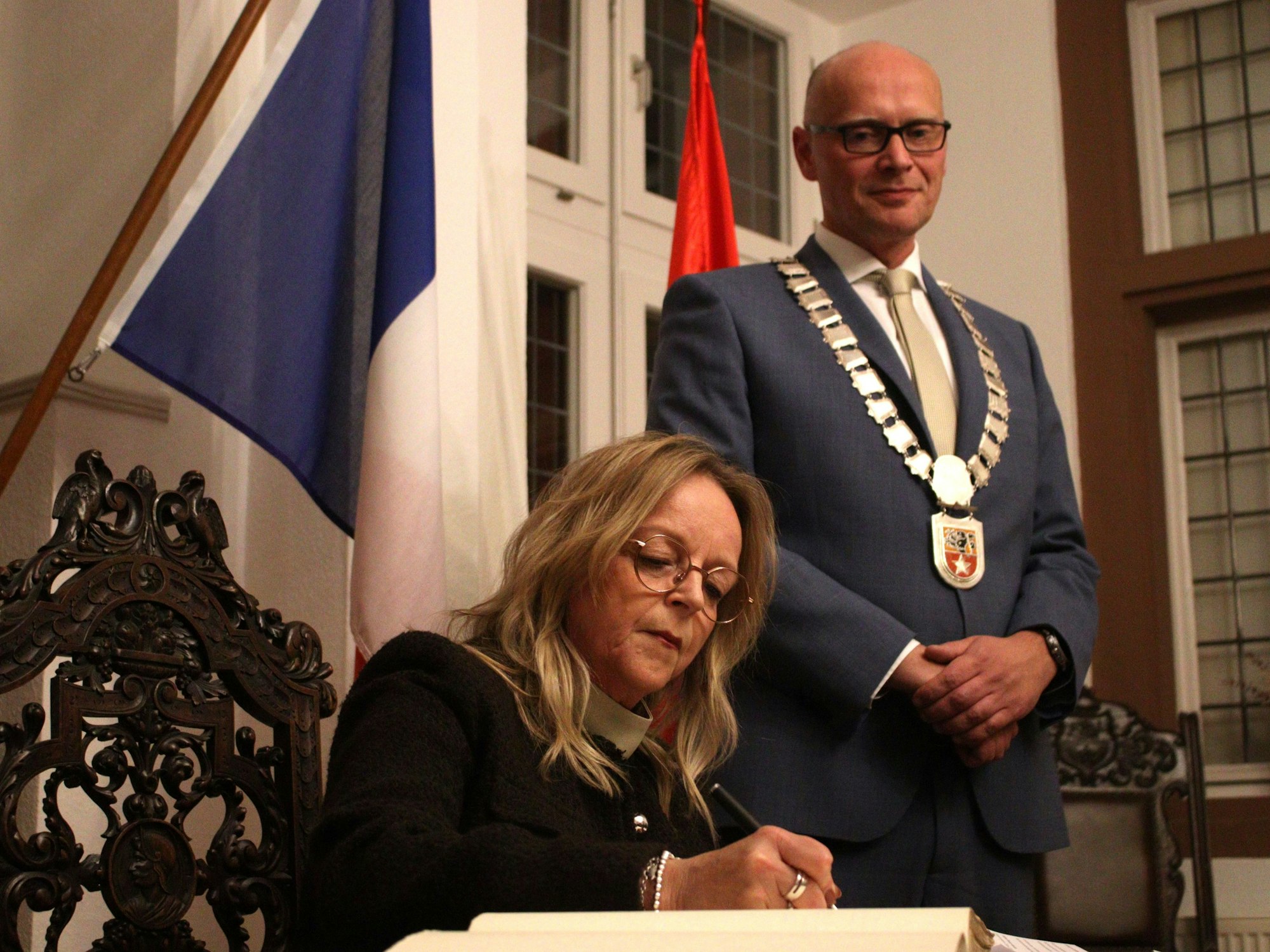 Sabine Preiser-Marian sitzt am Schreibtisch und schreibt ins Goldene Buch, Sebastian Glatzel steht daneben und schaut zu. Er trägt seine Amtskette.