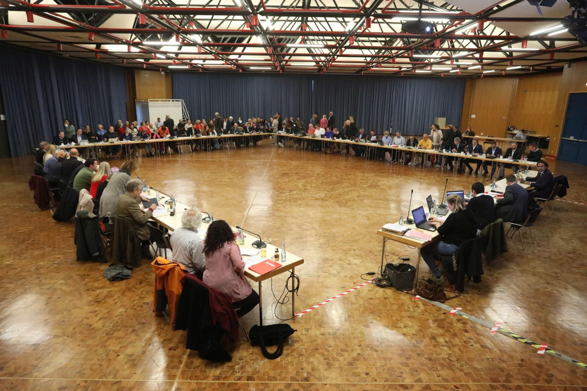 Viele Menschen sitzen in einer Aula an Tischen, die im Rechteck zusammengestellt sind.